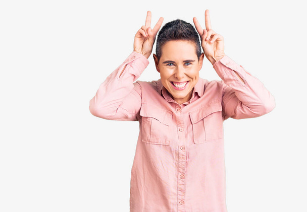 Νεαρή γυναίκα με κοντά μαλλιά φορώντας casual ρούχα που θέτουν αστείο και τρελό με τα δάχτυλα στο κεφάλι ως αυτιά λαγουδάκι, χαμογελώντας χαρούμενα  - Φωτογραφία, εικόνα