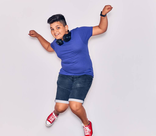 Adorable hispanic child boy smiling happy. Jumping with smile on face listening to music using headphones over isolated white background - Фото, изображение