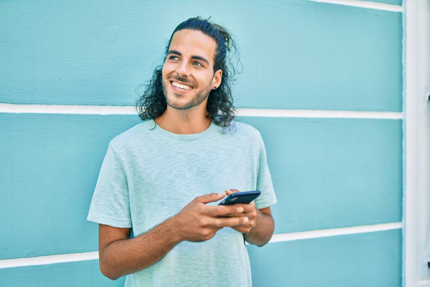 Młody Hiszpan uśmiecha się szczęśliwy korzystając ze smartfona opierającego się na ścianie - Zdjęcie, obraz