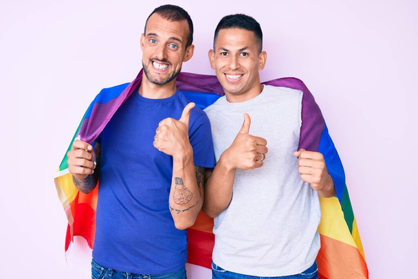 Молодая пара геев двое мужчин, держащих радугу lgbtq флаг вместе улыбаясь счастливый и позитивный, большой палец вверх делает отлично и знак одобрения  - Фото, изображение