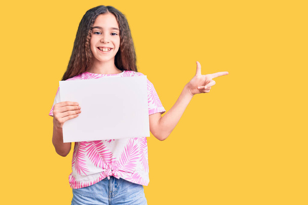 Linda niña hispana sosteniendo banner vacío en blanco sonriendo feliz señalando con la mano y el dedo hacia un lado  - Foto, Imagen
