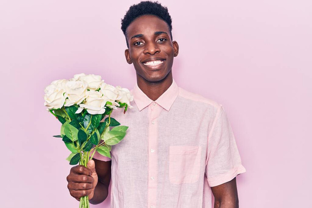 Fiatal afro-amerikai férfi kezében virágok látszó pozitív és boldog álló és mosolygó magabiztos mosollyal mutatja fogak  - Fotó, kép