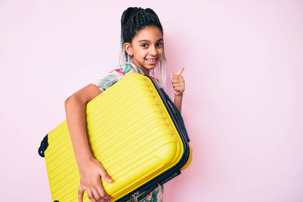 jong Afrikaans amerikaans meisje kind met vlechten houden koffer gaan op zomervakantie glimlachen gelukkig en positief, duim omhoog doen uitstekend en goedkeuring teken  - Foto, afbeelding