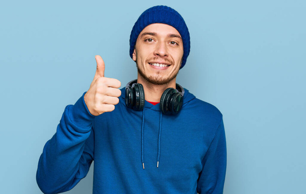 Hispanic young man wearing sweatshirt and wool hat smiling happy and positive, thumb up doing excellent and approval sign  - 写真・画像