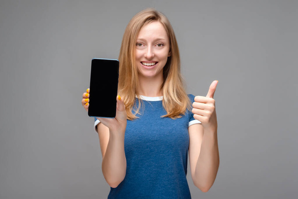 Fiatal gyönyörű szőke nő mutatja képernyő okostelefon felett elszigetelt háttér boldog nagy mosollyal csinál OK jel, hüvelykujj fel ujjaival, kiváló jel - Fotó, kép