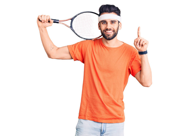 Young handsome man with beard playing tennis holding racket surprised with an idea or question pointing finger with happy face, number one  - Photo, Image
