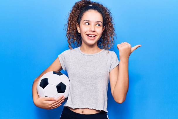 Hermosa niña con el pelo rizado sosteniendo pelota de fútbol apuntando pulgar hacia el lado sonriendo feliz con la boca abierta  - Foto, Imagen