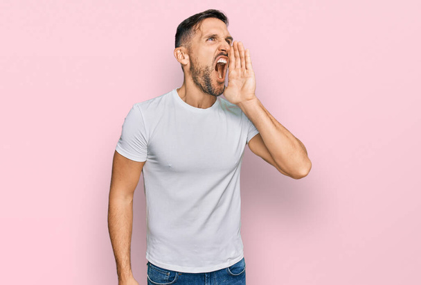 Handsome man with beard wearing casual white t shirt shouting and screaming loud to side with hand on mouth. communication concept.  - Photo, Image