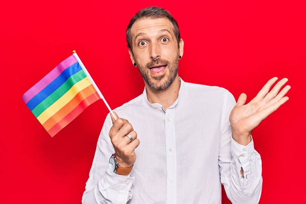 Jovem homem bonito segurando arco-íris lgbtq bandeira celebrando realização com sorriso feliz e expressão vencedora com mão levantada  - Foto, Imagem