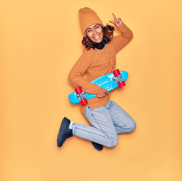 Young beautiful latin skater woman smiling happy. Jumping with smile on face holding skate doing victory sign over isolated yellow background - Photo, Image