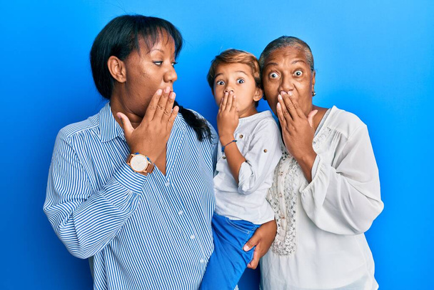 Hispanic family of grandmother, mother and son hugging together covering mouth with hand, shocked and afraid for mistake. surprised expression  - Zdjęcie, obraz