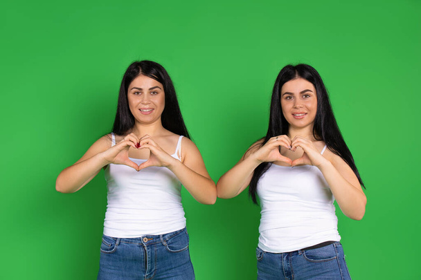 Twin sisters show a heart from their hands on a green background. St.Valentine's Day, charity and giving back. High quality photo - Foto, immagini