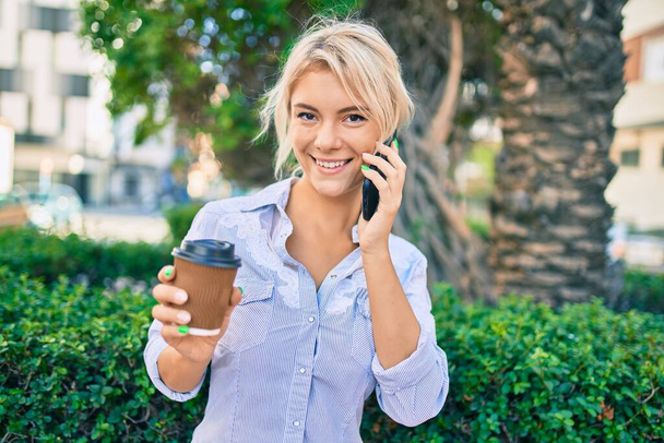 Junge blonde Frau lächelt glücklich mit dem Smartphone und trinkt Kaffee zum Mitnehmen in der Stadt. - Foto, Bild