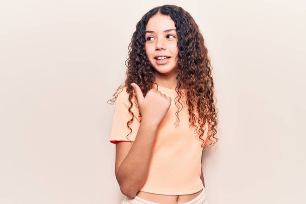 Beautiful kid girl with curly hair wearing casual clothes smiling with happy face looking and pointing to the side with thumb up.  - Foto, imagen