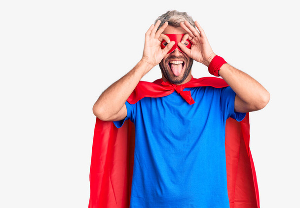 Young blond man wearing super hero custome doing ok gesture like binoculars sticking tongue out, eyes looking through fingers. crazy expression.  - Photo, Image