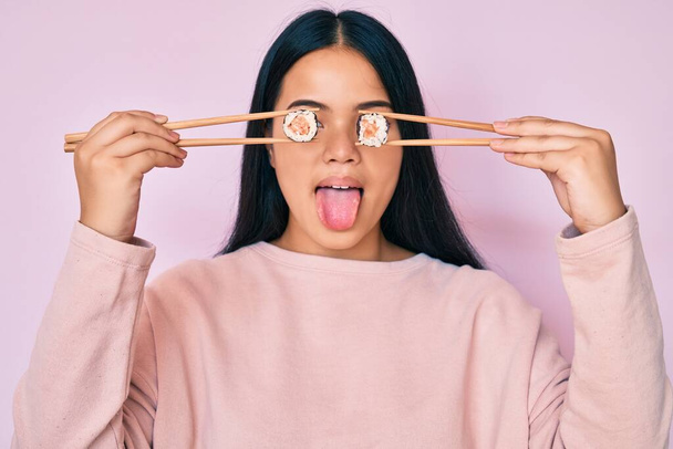 Young beautiful asian girl puting sushi on eyes using chopsticks sticking tongue out happy with funny expression.  - Photo, Image