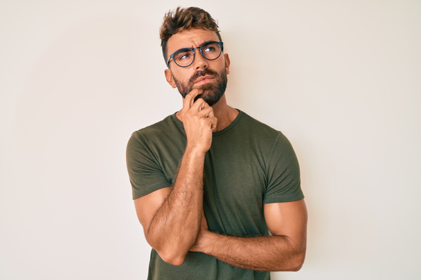 Mladý hispánec v neformálním oblečení a brýlích vážný obličej přemýšlející o otázce s rukou na bradě, přemýšlivý o matoucím nápadu  - Fotografie, Obrázek