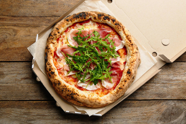 Sabrosa pizza con carne y rúcula en caja de cartón sobre mesa de madera, vista superior - Foto, imagen
