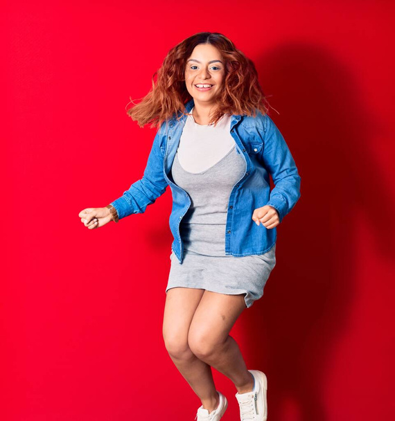 Νεαρή όμορφη Λατίνα γυναίκα που φοράει καθημερινά ρούχα χαμογελώντας ευτυχισμένη. Άλμα με χαμόγελο στο πρόσωπο κάνει χειρονομία νικητής με γροθιές πάνω από απομονωμένο κόκκινο φόντο - Φωτογραφία, εικόνα