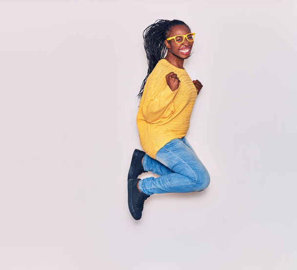 Young beautiful african american woman wearing casual clothes and glasses smiling happy. Jumping with smile on face doing winner gesture with fists up over isolated white background - Fotoğraf, Görsel