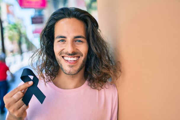 Joven hombre hispano sonriendo feliz sosteniendo amargado cinta negra apoyada en la pared. - Foto, Imagen