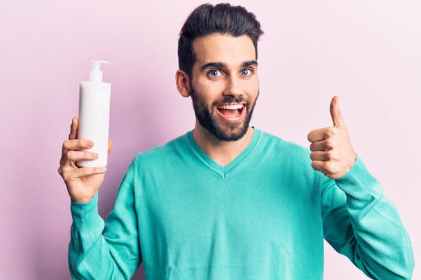 Young handsome man with beard holding bottle of cream smiling happy and positive, thumb up doing excellent and approval sign  - Fotó, kép