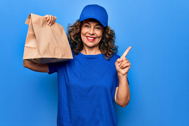 Middelbare leeftijd mooie levering vrouw holding leveren papieren zak met afhaalmaaltijden glimlachend gelukkig wijzend met hand en vinger naar de zijkant - Foto, afbeelding