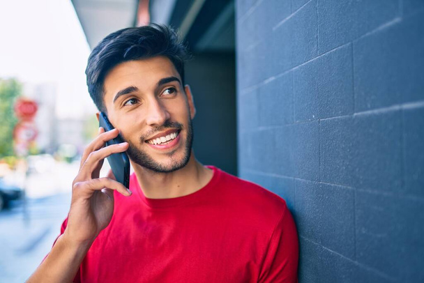 Genç Latin adam gülümsüyor. Şehirdeki akıllı telefondan konuşuyor.. - Fotoğraf, Görsel