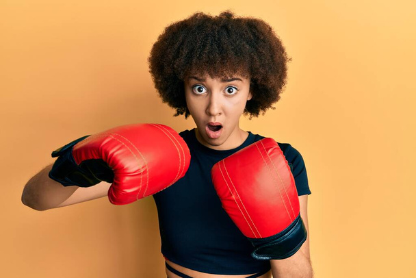 Jovem hispânica menina esportiva usando luvas de boxe em face de choque, olhando cético e sarcástico, surpreso com a boca aberta  - Foto, Imagem
