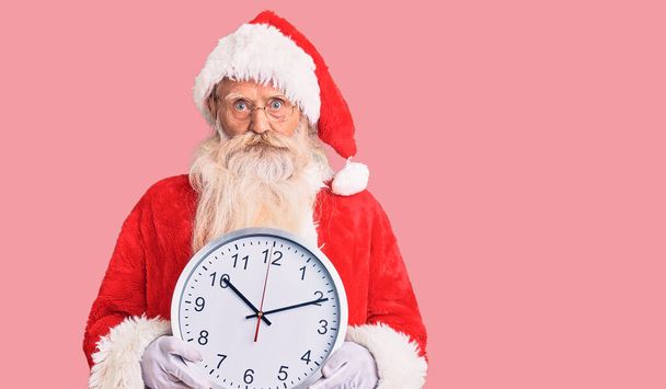 Старик с седыми волосами и длинной бородой в костюме Санта-Клауса держит часы, думая о настроении и трезвом выражении лица, выглядя уверенным в себе.  - Фото, изображение