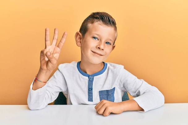 Entzückendes kaukasisches Kind in lässiger Kleidung, das auf dem Tisch sitzt und mit Finger Nummer drei nach oben zeigt, während es zuversichtlich und glücklich lächelt.  - Foto, Bild