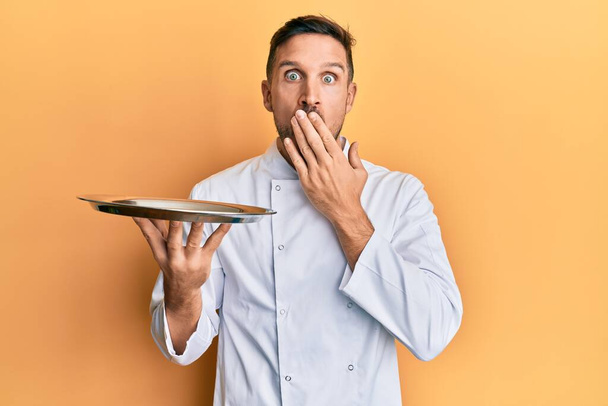 Pohledný muž s plnovousem v uniformě šéfkuchaře, držící stříbrný tác zakrývající ústa rukou, šokovaný a vystrašený z omylu. překvapený výraz  - Fotografie, Obrázek