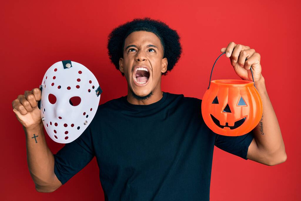African american man with afro hair wearing hockey mask and halloween pumpking angry and mad screaming frustrated and furious, shouting with anger looking up.  - Foto, imagen