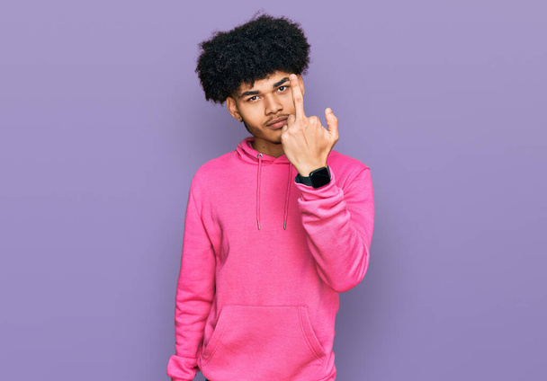 Young african american man with afro hair wearing casual pink sweatshirt showing middle finger, impolite and rude fuck off expression  - Photo, Image