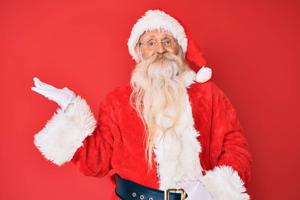 Alter älterer Mann mit grauen Haaren und langem Bart in traditioneller Weihnachtsmanntracht, fröhlich lächelnd und mit Handfläche in die Kamera zeigend.  - Foto, Bild