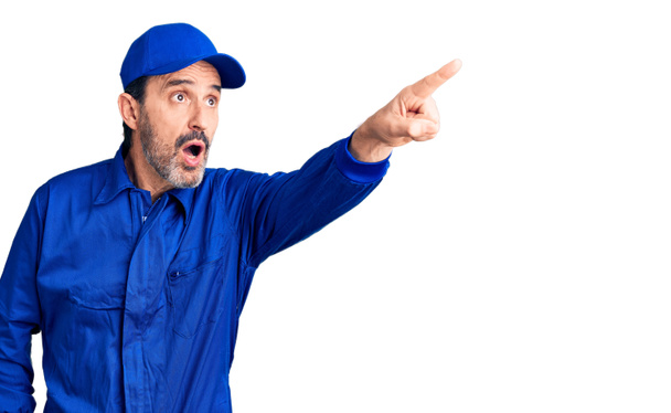 Middle age handsome man wearing mechanic uniform pointing with finger surprised ahead, open mouth amazed expression, something on the front  - 写真・画像
