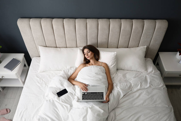 Tired woman with laptop and smartphone sleeping in bed at home, above view - Фото, изображение