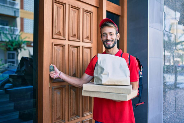 Kaukaski dostawca w czerwonym mundurze i plecaku dostawy uśmiechnięty szczęśliwy na zewnątrz trzymając pudełko pizzy - Zdjęcie, obraz