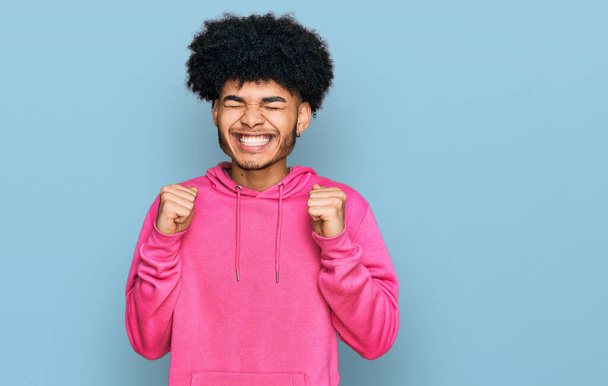 Fiatal afro-amerikai férfi afro haj visel alkalmi rózsaszín pulóver izgatott a siker karját, és csukott szemmel ünnepli győzelem mosolyogva. győztes koncepció.  - Fotó, kép