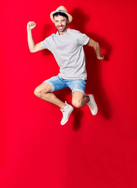 Young handsome hispanic man wearing casual clothes and summer hat smiling happy. Jumping with smile on face celebrating with fists up over isolated red background. - Valokuva, kuva