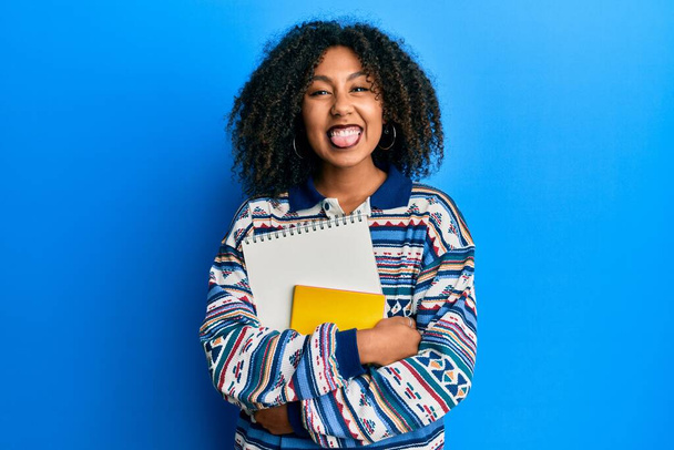 Hermosa mujer afroamericana con cabello afro sosteniendo libros sacando la lengua feliz con expresión divertida.  - Foto, imagen