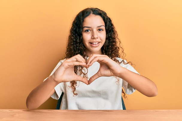 Teenager hispanische Mädchen in legerer Kleidung sitzen auf dem Tisch und lächeln verliebt und tun Herzsymbolform mit den Händen. Romantisches Konzept.  - Foto, Bild