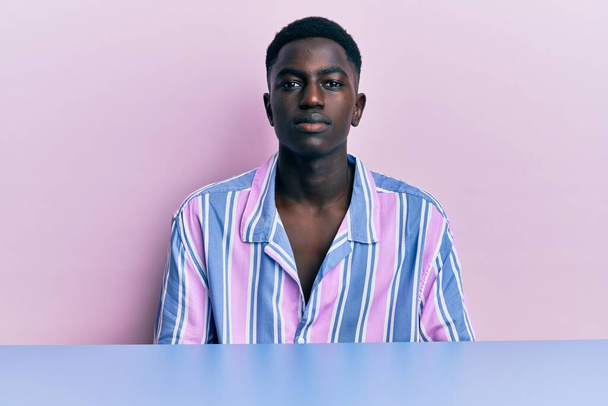 Young african american man wearing casual clothes sitting on the table relaxed with serious expression on face. simple and natural looking at the camera.  - Foto, imagen