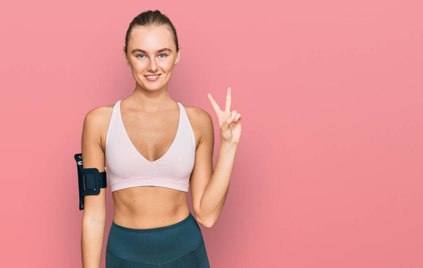 Beautiful young blonde woman wearing sportswear and arm band smiling with happy face winking at the camera doing victory sign. number two.  - Photo, Image
