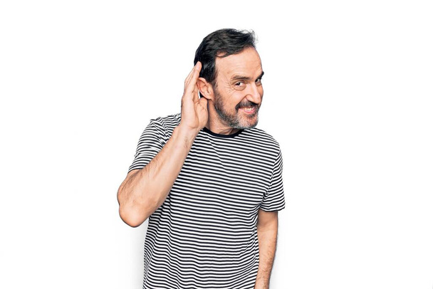Мужчина средних лет в полосатой футболке стоял на изолированном белом фоне улыбаясь рукой над ухом слушая и слушая слухи или сплетни. Концепция глухоты
. - Фото, изображение