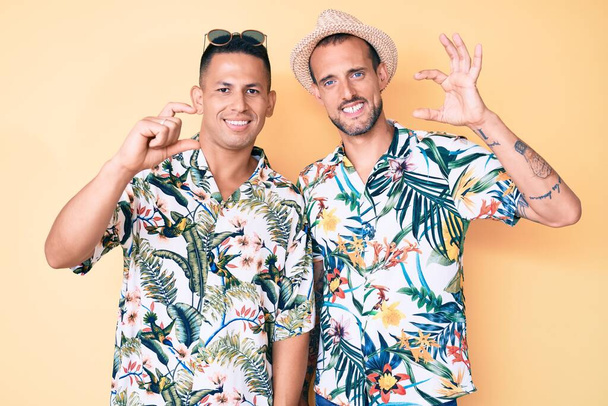 Joven pareja gay de dos hombres con sombrero de verano y camisa hawaiana sonriente y un gesto seguro con la mano haciendo un cartel de tamaño pequeño con los dedos mirando y la cámara. concepto de medida.  - Foto, Imagen