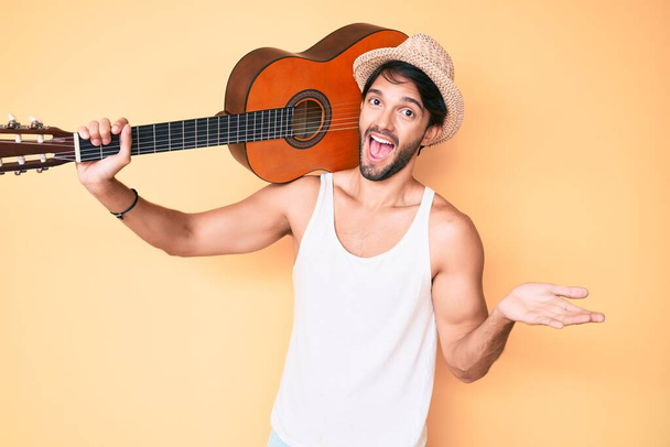 Hombre hispano guapo con guitarra clásica celebrando logro con sonrisa feliz y expresión ganadora con la mano levantada  - Foto, imagen