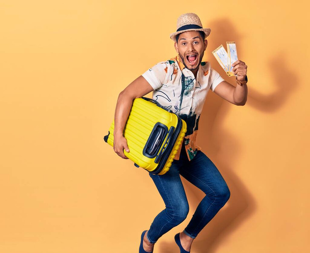 Junger gutaussehender lateinischer Mann im Urlaub in Sommerkleidung, der glücklich lächelt. Springen mit einem Lächeln im Gesicht mit Handtasche und Flugzeugbordkarte über isoliertem Hintergrund - Foto, Bild