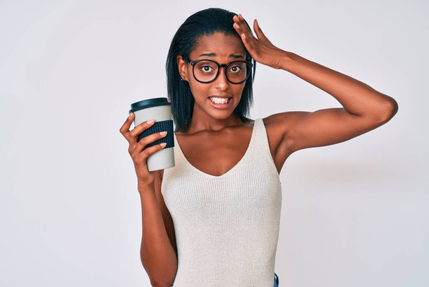 Mujer afroamericana joven sosteniendo tomar café estresado y frustrado con la mano en la cabeza, cara sorprendida y enojada  - Foto, Imagen