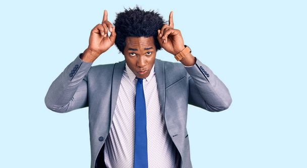 Bell'uomo afro-americano con i capelli afro che indossa una giacca da lavoro facendo un gesto divertente con un dito sopra la testa come corna di toro  - Foto, immagini